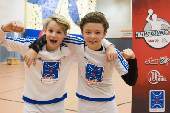 Zwei lächelnde Jungen in passenden weißen Hemden mit blauen Streifen und Handball-Camp-Logos posieren mit den Armen umeinander in einer Turnhalle, wobei ein Junge seinen Arm anspannt. Neben ihnen steht ein rotes Banner mit Logos.