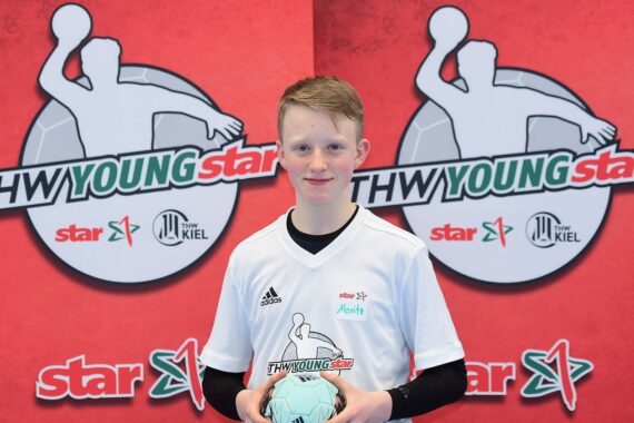 Ein Jugendlicher in einem weißen Sporttrikot hält einen hellblauen Handball in der Hand und steht vor einem roten Hintergrund mit THW-Youngstar-Logos und der Grafik einer Person, die einen Ball wirft.