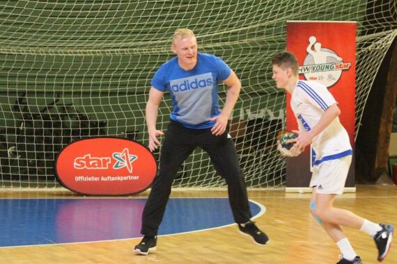 Zwei Männer spielen in einer Halle Handball; einer in einem blauen Adidas-Trikot und einer schwarzen Hose verteidigt, während der andere in einer weißen Uniform mit dem Ball rennt, mit einem Netz und Sponsorenbannern im Hintergrund.