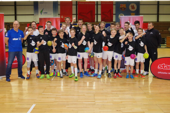 Eine Jugendhandballmannschaft in passenden Trikots posiert energisch in einer Turnhalle mit ihren Trainern, die bunte Bälle halten. Im Hintergrund sind Banner und Sponsorenlogos auf dem Holzplatz zu sehen.