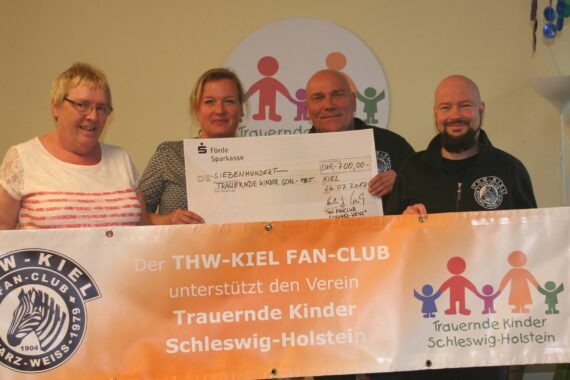 Vier Personen lächeln, während sie einen großen Spendenscheck und ein Transparent mit der Aufschrift THW-Kiel Fan-Club unterstützt den Verein Trauernde Kinder Schleswig-Holstein" halten. Hinter ihnen sind ein Logo und ein Wandschild der Organisation zu sehen.
