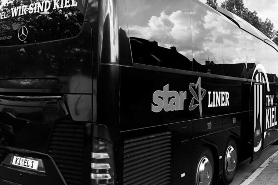 Ein schwarzer Bus mit den Logos von star LINER und THW KIEL ist auf einer Straße geparkt. Der Bus hat getönte Scheiben, in denen sich Wolken und Gebäude spiegeln. Auf dem Nummernschild steht K1 EL 1.