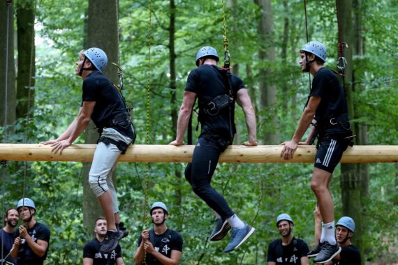Drei Personen, die Helme und Klettergurte tragen, balancieren auf einem aufgehängten Holzstamm in einem Wald, während mehrere andere Personen, die ebenfalls Helme tragen, während einer Teambildungsaktivität im Freien vom Boden aus zusehen.