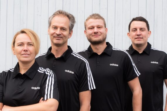 Vier Personen stehen in einer Reihe vor einer hellen Holzwand, alle tragen passende schwarze Adidas-Shirts mit weißen Streifen an den Schultern. Sie blicken in die Kamera und lächeln.