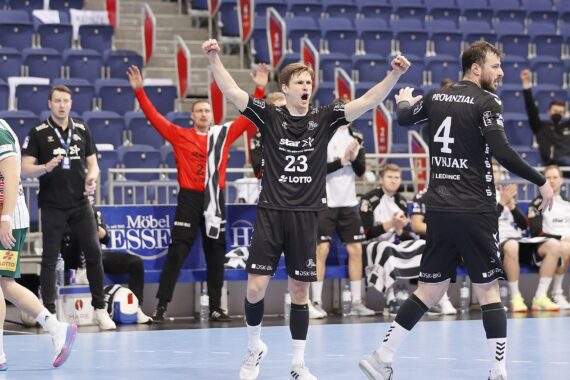 Zwei männliche Handballspieler in schwarzen Trikots feiern auf dem Spielfeld, indem sie die Arme zum Sieg erheben, während ihre Mannschaftskameraden und Trainer im Hintergrund jubeln. Ein gegnerischer Spieler in einem grün-weißen Trikot schaut zu.