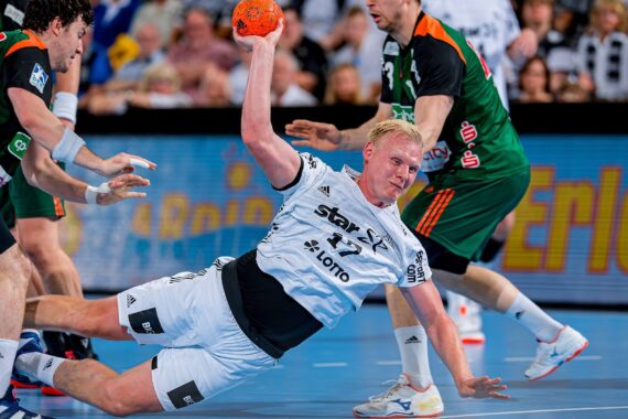 Ein männlicher Handballspieler in einem weißen Trikot fällt zu Boden, während er einen orangefarbenen Ball in der Hand hält und die anderen Spieler um ihn herum um den Ballbesitz kämpfen.