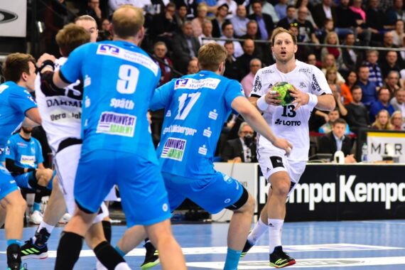 Ein Handballspieler in einem weißen Trikot hält den Ball und bereitet sich auf einen Spielzug vor, während drei Spieler in blauen Trikots verteidigen. Die Szene spielt sich in einer Halle ab, mit Zuschauern im Hintergrund.