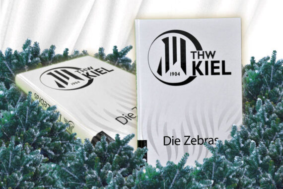 Zwei weiße Bücher mit dem Logo des THW Kiel und dem Titel "Die Zebras" stehen zwischen grünen Tannenzweigen, vor einem weißen, drapierten Stoffhintergrund.