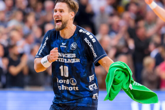 Ein männlicher Handballspieler in einem blauen Trikot mit der Nummer 10 jubelt energisch, während er während eines Spiels ein grünes Handtuch in der Hand hält. Im unscharfen Hintergrund ist eine jubelnde Menge zu sehen.
