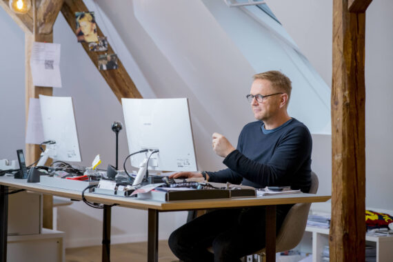 Ein Mann mit Brille sitzt an einem Schreibtisch in einem modernen Büro und arbeitet an einem Computer. Der Arbeitsbereich ist mit Holzbalken, Papieren und Büromaterial ausgestattet, und durch ein großes Fenster fällt natürliches Licht ein.