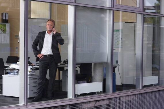 Ein Mann im Geschäftsanzug steht in einem Büro, telefoniert mit einem Handy und schaut aus dem großen Fenster. Auf einem Schreibtisch liegen Stapel von Papieren und im Inneren sind Büromöbel zu sehen.