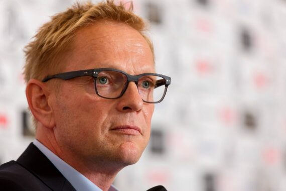 Ein Mann mit kurzen blonden Haaren und Brille, der einen Anzug trägt, schaut leicht zur Seite, während er in ein Mikrofon spricht. Der Hintergrund ist unscharf mit weißen und schwarzen Mustern.
