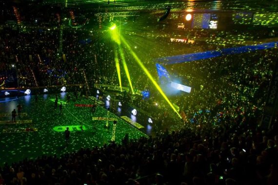 Eine große, vollbesetzte Halle mit farbenfrohen grünen und gelben Laserlichtern und Scheinwerfern, die das Publikum und die Bühne anstrahlen und eine festliche, energiegeladene Atmosphäre während einer Veranstaltung oder eines Konzerts schaffen.