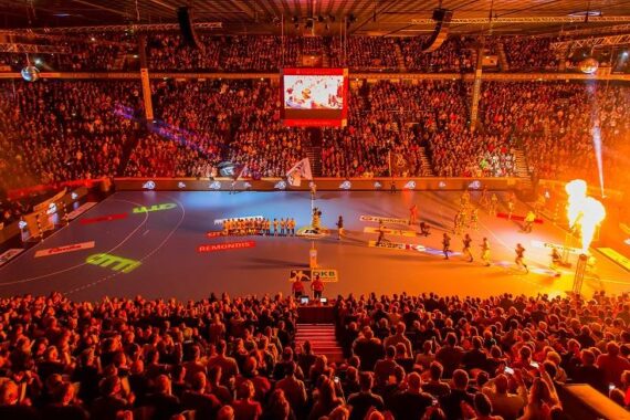 Eine große, überfüllte Halle mit hellen Lichtern und Feuereffekten, in der zwei Sportmannschaften vor einem Handballspiel auf einem blauen Spielfeld aufgereiht stehen, während die Zuschauer von der Tribüne aus zusehen.