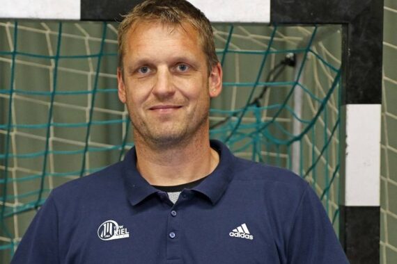 Ein Mann in einem marineblauen Sport-Poloshirt steht vor einem Tornetz in einer Sporthalle und lächelt in die Kamera.