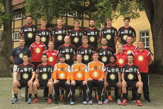 Eine Herren-Handballmannschaft stellt sich im Freien in vier Reihen auf und trägt schwarze und orangefarbene Trikots mit Sonnenlogos. Trainer und Betreuer in Rot stehen an den Seiten. Im Hintergrund sind Bäume und Gebäude zu sehen.