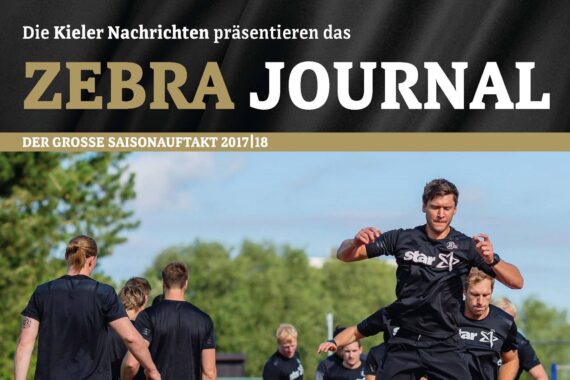 Eine Gruppe von Fußballspielern in schwarzer Trainingskleidung übt im Freien unter blauem Himmel. Am oberen Rand des Bildes steht in großen Buchstaben der Text Zebra Journal.