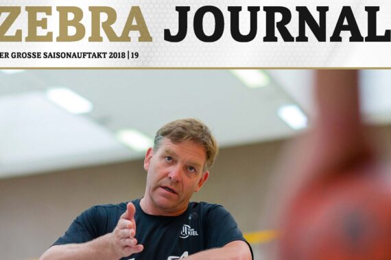 Ein Mann in einem dunklen Hemd gestikuliert mit seinen Händen, als wolle er etwas erklären, auf dem Cover einer Zeitschrift mit dem Titel Zebra Journal. Im Hintergrund ist ein Innenraum mit hellen Lichtern zu sehen.