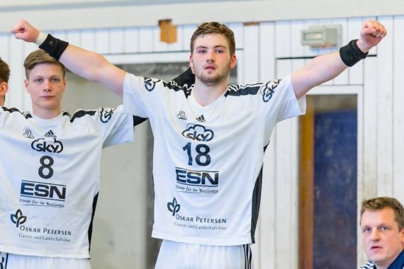 Ein männlicher Handballspieler in einem weißen Trikot mit der Nummer 18 steht mit erhobenen Armen da und jubelt. Ein anderer Spieler mit der Trikotnummer 8 steht neben ihm. Beide befinden sich in einer Halle, ein Trainer oder Offizieller ist in der Nähe.
