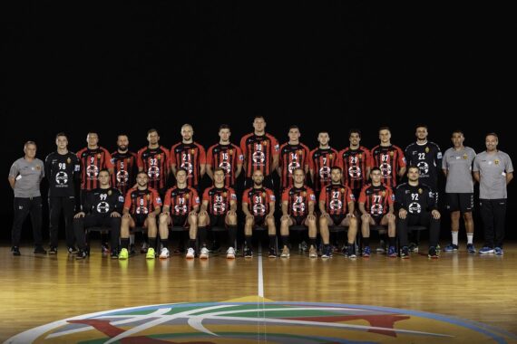 Eine Männerhandballmannschaft posiert in einem Innenraum auf einem Spielfeld, in passenden schwarzen und roten Trikots, mit Trainern und Mitarbeitern in grauer Kleidung, die auf beiden Seiten vor einem schwarzen Hintergrund stehen.
