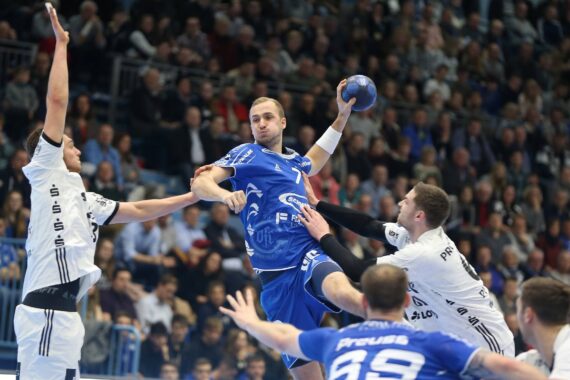 Ein männlicher Handballspieler in einem blauen Trikot springt, um zu schießen, während er während eines Spiels von drei Gegnern in weißen Trikots geblockt wird, während ein großes Publikum im Hintergrund zusieht.
