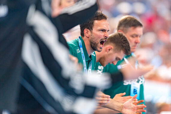 Eine Gruppe von Männern in grünen Sportuniformen feuert ihre Mannschaft bei einem Sportereignis leidenschaftlich an, wobei ein Mann in der Mitte schreit und klatscht. Die Atmosphäre ist intensiv und energiegeladen.