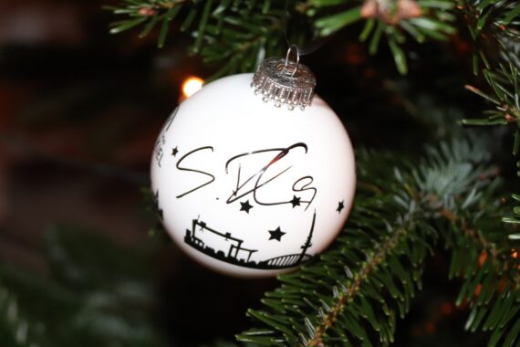 Ein weißes Weihnachtsornament mit schwarzem Stadtbild und Sternenmotiven hängt an einem grünen Tannenzweig, mit warmen Lichtern im Hintergrund.