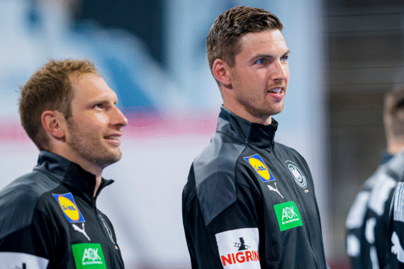 Zwei männliche Sportler in schwarzen Sportjacken mit Sponsorenlogos stehen nebeneinander, schauen nach vorne und lächeln leicht. Der Hintergrund ist unscharf, um die beiden Personen in den Vordergrund zu rücken.