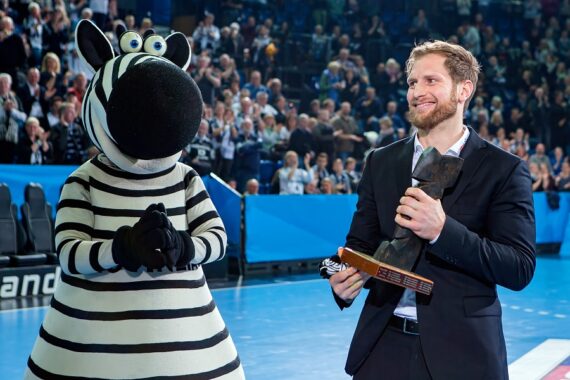 Ein Mann im Anzug, der einen Pokal hält, steht neben einer Person in einem Zebra-Maskottchenkostüm auf dem Boden einer Sportarena, mit einer jubelnden Menge im Hintergrund.