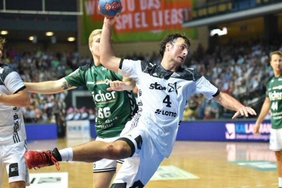 Ein Handballspieler im weißen Trikot springt, während er den Ball hält und sich zum Wurf vorbereitet, während ein Verteidiger im grünen Trikot versucht, ihn während eines Spiels in einer Halle zu blockieren.