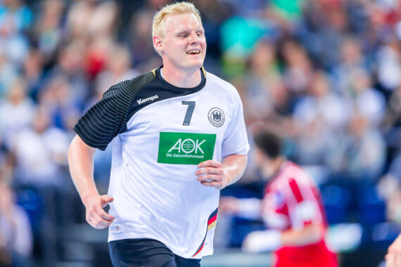 Ein männlicher Handballspieler, der ein weißes Trikot mit der Nummer 7 und einer deutschen Flagge trägt, lächelt, während er auf das Spielfeld läuft. Im Hintergrund ist ein weiterer Spieler in Rot und eine Zuschauermenge zu sehen.