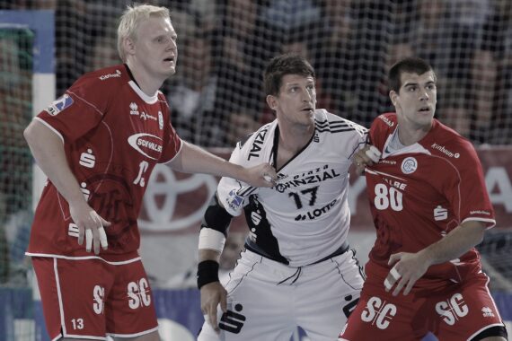 Drei männliche Handballspieler kämpfen während eines Spiels um die Position; zwei Spieler in Rot verteidigen gegen einen Spieler in Weiß, während im Hintergrund ein Tor zu sehen ist und die Zuschauer von der Tribüne aus zusehen.