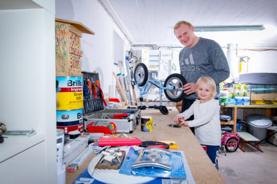 Ein lächelnder Erwachsener und ein Kind arbeiten zusammen in einer hellen Garagenwerkstatt und montieren ein kleines Fahrrad auf einer unübersichtlichen Werkbank, die mit Werkzeugen und verschiedenen Materialien gefüllt ist.