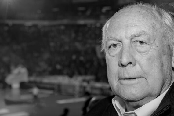 Ein älterer Mann mit schütterem weißen Haar und ernster Miene blickt in die Kamera. Der Hintergrund scheint eine Sporthalle mit unscharfen Zuschauern und einem Spielfeld zu sein. Das Bild ist in schwarz-weiß gehalten.