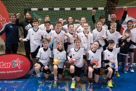 Eine Jugendhandballmannschaft in weißen Trikots posiert aufgeregt vor einem Tornetz mit ihren Trainern, von denen einige Handbälle halten. Alle lächeln und machen feierliche Gesten. Auf der linken Seite ist ein Banner eines Starsponsors zu sehen.