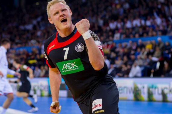 Ein männlicher Handballspieler in einem schwarzen Trikot mit der Nummer 7 ballt die Faust und jubelt auf dem Spielfeld, während im Hintergrund eine verschwommene Menschenmenge und andere Spieler zu sehen sind.