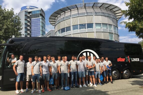Eine Gruppe von Menschen in passenden hellgrauen Hemden und Shorts steht lächelnd vor einem großen schwarzen Reisebus mit der Aufschrift star LINER, mit modernen Bürogebäuden und Bäumen im Hintergrund an einem sonnigen Tag.