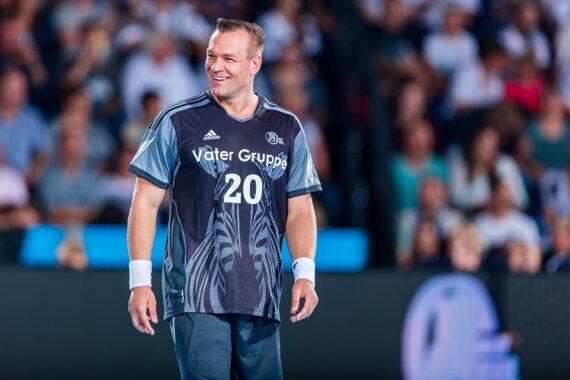 Ein lächelnder männlicher Handballspieler, der ein dunkles Trikot mit der Nummer 20 trägt, steht auf dem Spielfeld, mit einer verschwommenen Menschenmenge im Hintergrund.