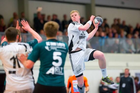 Ein männlicher Handballspieler in einem weißen Trikot springt in der Luft und bereitet sich auf den Wurf vor, während die gegnerischen Spieler in Grün den Ball abwehren. Eine verschwommene Menschenmenge beobachtet das intensive Spiel im Hintergrund.