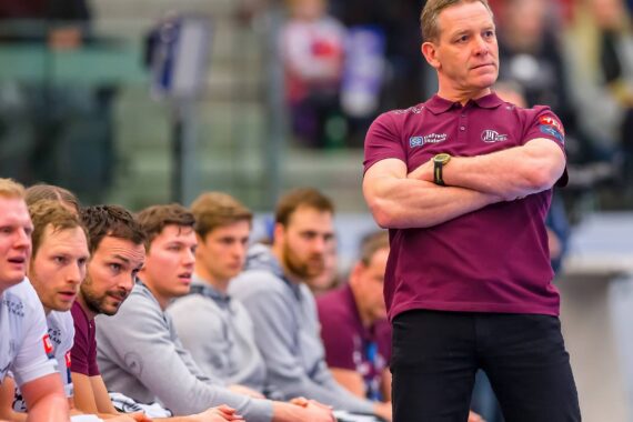Ein Trainer in einem kastanienbraunen Hemd steht mit verschränkten Armen vor einer Reihe von sitzenden Sportlern, die während eines Hallensportspiels aufmerksam zusehen. Im Hintergrund ist ein unscharfes Publikum zu sehen.