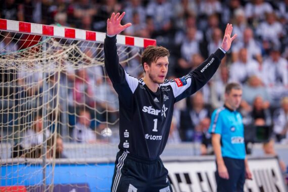 Ein männlicher Handballtorwart in dunkler Uniform steht mit erhobenen Armen vor dem Tor, bereit zur Abwehr. Im Hintergrund sind Zuschauer und ein Schiedsrichter in Blau zu sehen.
