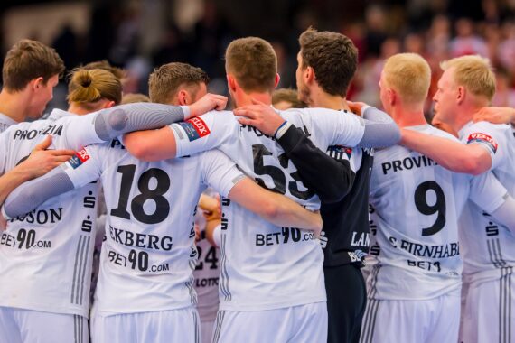 Eine Gruppe männlicher Handballspieler in weißen Trikots steht zusammen und legt sich gegenseitig die Arme um die Schultern, um Teamwork und Einigkeit vor einem Spiel zu demonstrieren. Ihre Trikotnummern und Sponsorenlogos sind sichtbar.