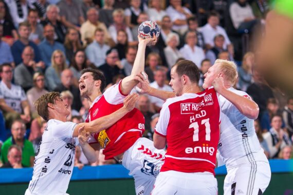 Ein Handballspieler in einem roten Trikot springt und versucht zu schießen, während er von drei Gegnern in weißen Trikots verteidigt wird, während die Zuschauer im Hintergrund aufmerksam zusehen.