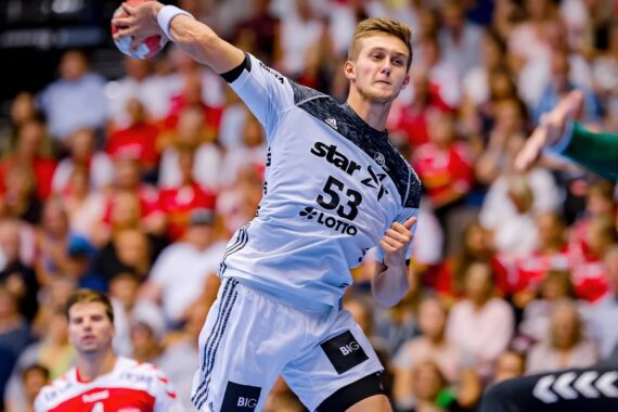 Ein männlicher Handballspieler, der ein weißes Trikot mit der Nummer 53 trägt, bereitet sich während eines Spiels auf den Wurf vor, während im Hintergrund unscharfe Zuschauer und Gegner in roten Trikots zu sehen sind.