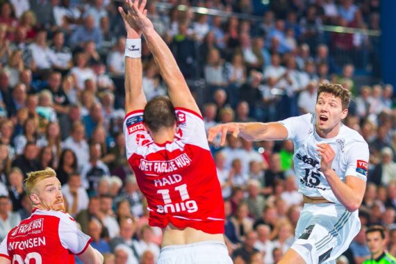 Ein Handballspieler in einem weißen Trikot springt, um den Ball an zwei Verteidigern in roten Trikots vorbei zu schießen, während im Hintergrund eine Menge Zuschauer zusieht.