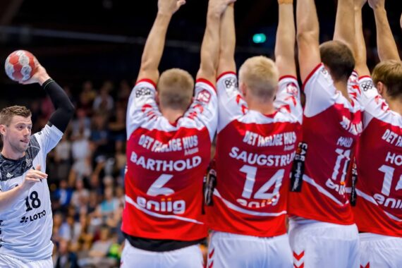 Ein Handballspieler in einem weißen Trikot bereitet sich auf den Wurf vor, während vier Verteidiger in rot-weißen Trikots mit erhobenen Armen aufspringen, um den Wurf zu blockieren.