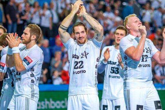 Eine Herren-Handballmannschaft in weißen Trikots steht gemeinsam auf dem Spielfeld, klatscht und feiert vor einem Publikum. Der Spieler in der Mitte hat die Hände über den Kopf erhoben und applaudiert mit einem Lächeln.