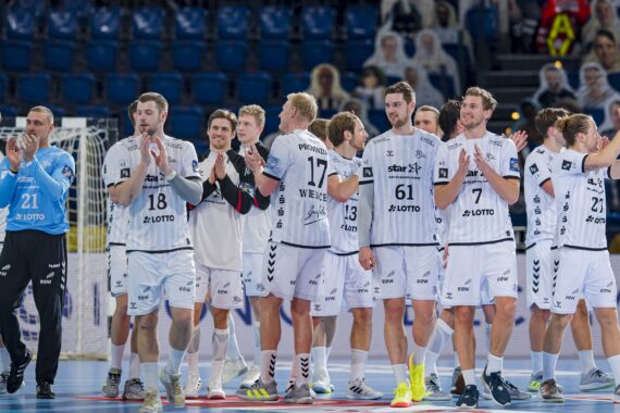 Eine Männerhandballmannschaft in weißen Trikots steht zusammen auf einem Hallenplatz, klatscht und lächelt, während im Hintergrund leere blaue Sitze und einige Zuschauer zu sehen sind.