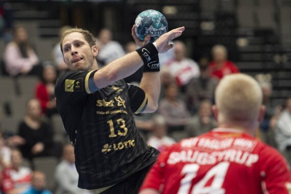Ein Handballspieler im schwarzen Trikot, Nummer 13, bereitet sich auf den Wurf vor, während er einem Abwehrspieler im roten Trikot, Nummer 14, während eines Spiels in einer Halle mit Zuschauern im Hintergrund gegenübersteht.