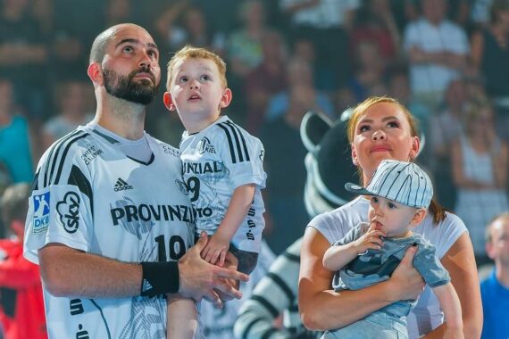 Ein Mann und eine Frau, die jeweils ein kleines Kind im Arm halten, stehen zusammen in einer Sporthalle. Alle vier blicken interessiert nach oben. Der Mann und das ältere Kind tragen passende Sporttrikots, die Frau hält ein Baby mit einer gestreiften Mütze.